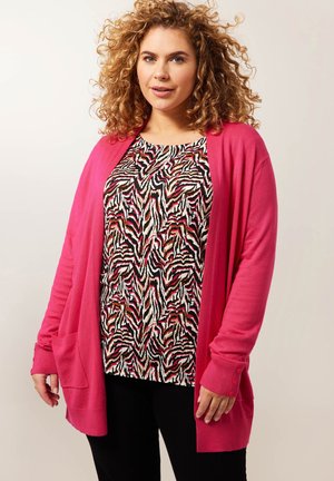 Lange roze cardigan met zakken, gedragen over een top met zwart-wit patroon en rode en roze accenten. Zachte textuur, losse pasvorm.