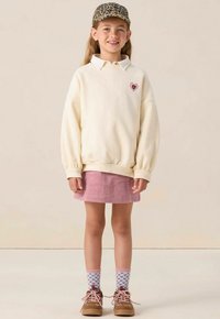 Crème sweatshirt met oversized mouwen en hartontwerp, roze corduroy rok, gestreepte sokken en bruine schoenen. Accent met een pet van luipaardprint.