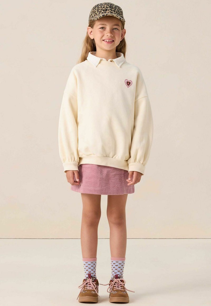 Crème sweatshirt met oversized mouwen en hartontwerp, roze corduroy rok, gestreepte sokken en bruine schoenen. Accent met een pet van luipaardprint.