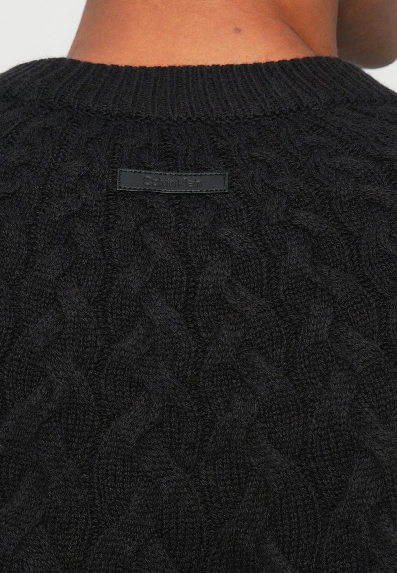 Maglione nero lavorato a maglia con motivo a trecce, caratterizzato da una scollatura a coste e un'etichetta rettangolare nera con la scritta "Calvin Klein" sul retro.