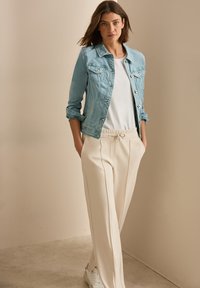 Chaqueta de mezclilla azul claro sobre una camiseta blanca, combinada con pantalones de chándal crema con pliegues y zapatillas blancas. Texturas suaves y ajuste relajado.