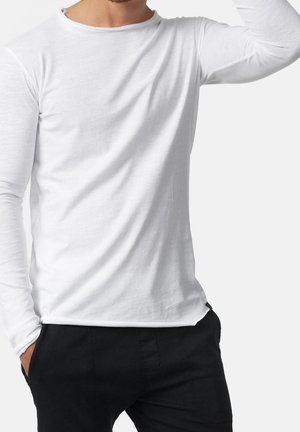 Homme portant une chemise blanche unie à manches longues et un pantalon noir, une main dans la poche et l'autre levée près de la tête.