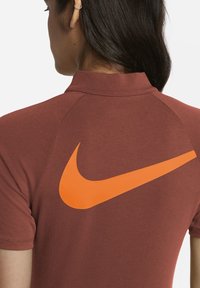 Brązowa sportowa koszulka polo z wysokim kołnierzem i pomarańczowym logo Nike na plecach. Gładki materiał z dopasowanym krojem i raglanowymi rękawami.