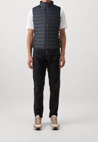 Svart pufferväst med horisontell stygn, hög krage och dragkedja fram. Kombinerad med svarta jeans och tan- och vita sneakers.