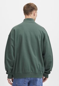 Sudadera de gran tamaño en verde oscuro, con un cuello alto y puños acanalados. Material de mezcla de algodón suave. Diseño minimalista, sin logotipos visibles.