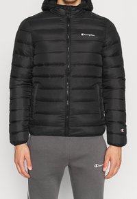 Chaqueta de abrigo negra con capucha, con acolchado horizontal, cierre de cremallera, bolsillos laterales y un pequeño logo blanco en el pecho y la manga.