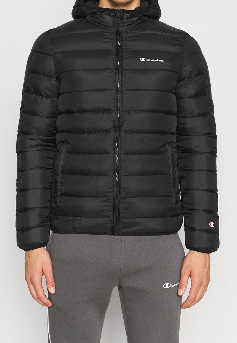 Chaqueta de abrigo negra con capucha, con acolchado horizontal, cierre de cremallera, bolsillos laterales y un pequeño logo blanco en el pecho y la manga.