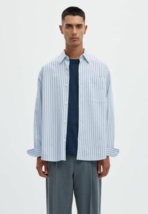 Jeune homme aux cheveux courts et foncés porte une chemise blanche ample à fines rayures verticales par-dessus un t-shirt bleu foncé et un pantalon gris.