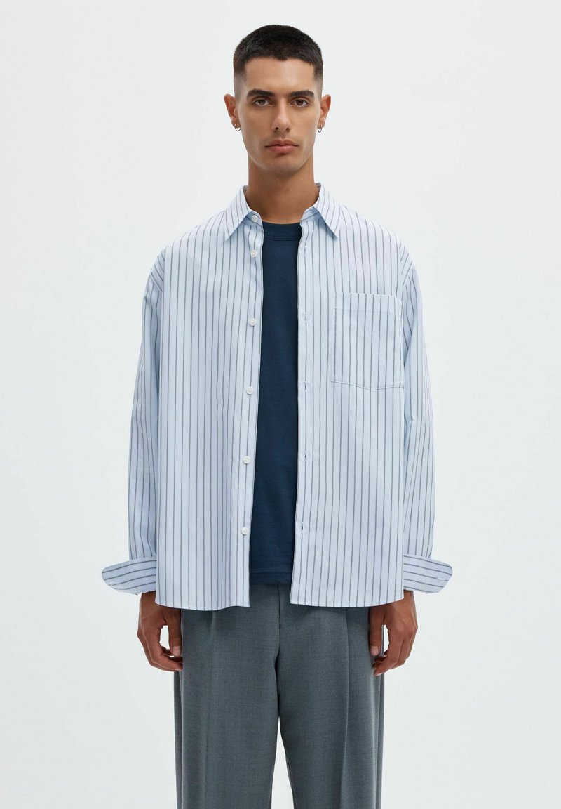 Jeune homme aux cheveux courts et foncés porte une chemise blanche ample à fines rayures verticales par-dessus un t-shirt bleu foncé et un pantalon gris.
