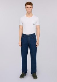 Camisa blanca de manga corta con un gráfico en el pecho, combinada con jeans rectos de pierna oscura y zapatillas verdes.