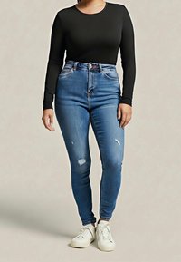 Personne portant un haut noir à manches longues, un jean skinny bleu taille haute avec des patchs usés, et des baskets blanches, debout sur un fond clair.