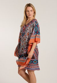 Monivärinen kaftan, jossa on geometrisia kuvioita mustassa, oranssissa ja turkoosissa. V-pääaukko ja leveät hihat, pehmeä kankaan rakenne.