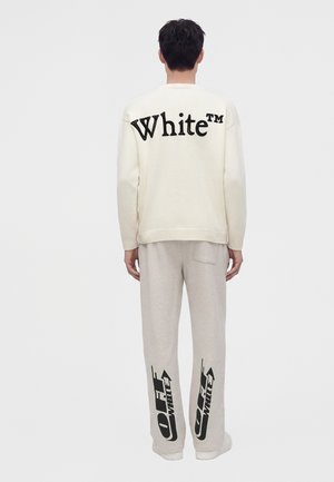 Personne vue de dos portant un pull blanc avec le texte "White" et un pantalon clair avec des logos noirs "Off-White" sur le bas des jambes.
