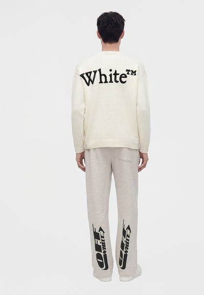 Personne vue de dos portant un pull blanc avec le texte "White" et un pantalon clair avec des logos noirs "Off-White" sur le bas des jambes.
