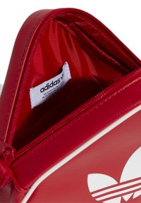 Pochette rossa con zip realizzata in materiale sintetico. L'interno presenta una fodera rossa liscia e un'etichetta adidas, con accenti bianchi all'esterno.