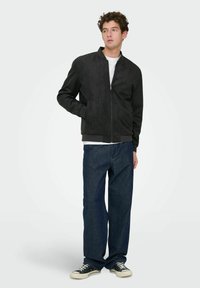 Jeune homme portant une veste noire zippée, une chemise blanche, un jean bleu ample et des baskets noires, debout avec une main dans la poche, regardant vers la droite.