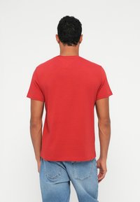 Camiseta corta roja de manga corta con cuello redondo, hecha de algodón suave. La vista trasera muestra un diseño limpio, sin estampados y con un dobladillo modesto. Jeans de denim complementan.