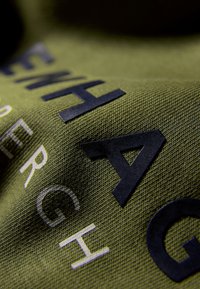 Olivengrön tyg med strukturerad väv, som har djärva svarta bokstäver i varierande storlekar, vilket skapar en lager-på-lager visuell effekt.