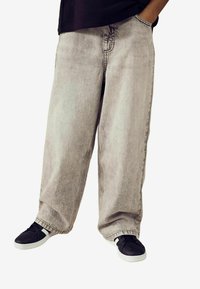 Helle graue weit geschnittene Jeans mit verwehtem Effekt, ausgestattet mit seitlichen Taschen und einem lockeren Schnitt. Kombiniert mit schwarzen Turnschuhen und einem schwarzen Hemd.