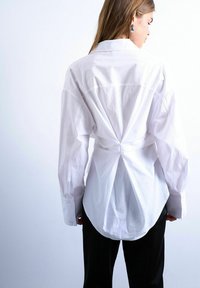 Femme aux cheveux longs portant une chemise blanche à boutons oversize avec un détail froncé à l'arrière et un pantalon noir, de dos sur un fond blanc.
