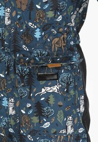 Sininen kangas, jossa on villieläinkuvio, mukaan lukien sudet, kanit, hirvet, tiikerit, oravat, puut, lehdet ja tammenterhot, taskun ympärillä, jossa on läppä ja tarranauha.