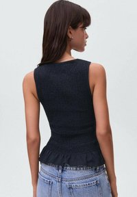 Model v črni smock bluzi in svetlih denim kratkih hlačah, obrnjena stran od nevtralne ozadja. Lase ima spuščene, vidne so obroče.