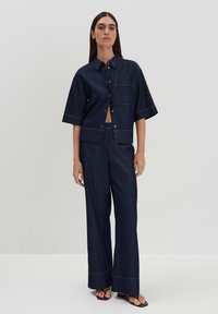 Navy denim shirt with short sleeves and front pocket, paired with wide-leg navy denim pants, both featuring contrasting yellow stitching.