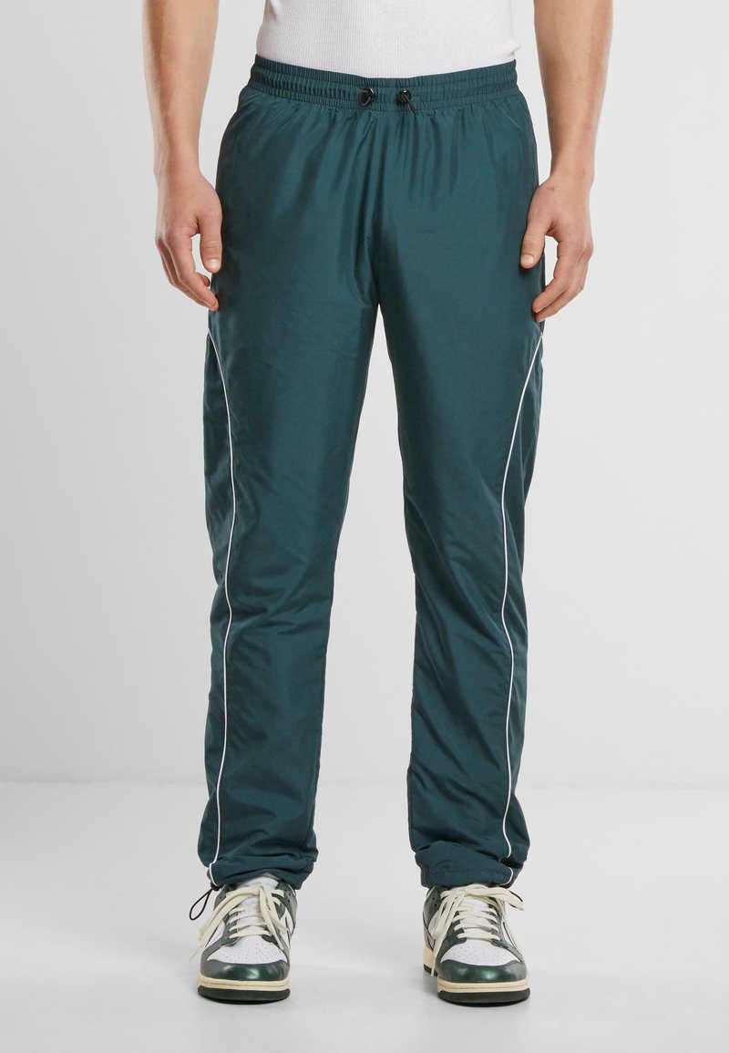 Pantalones de chándal ligeros en color teal con cintura elástica, cordón, ribete blanco en los costados y corte tapered en los tobillos. Combinados con zapatillas.