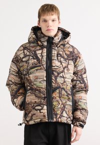 Polstret jakke med camouflage mønster i jordfarver, lynlås foran, snoretræk i bunden og hætte med justerbare snore.