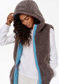 Hnědá fleecová vesta s kapucí, se zářivě modrým předním zipem a růžovými detaily. Vesta má strukturovaný, útulný vzhled.