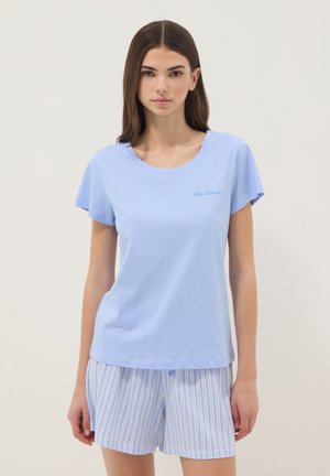 Young woman wearing light blue short-sleeve shirt and blue-and-white striped shorts, standing against a plain light background.