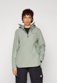Giacca impermeabile verde chiaro con zip frontale, cappuccio regolabile e tasche laterali, indossata sopra un top nero e pantaloni cargo scuri.