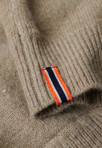 Beżowa dzianina o prążkowanej fakturze; posiada metkę w pomarańczowe, granatowe i białe paski. Miękki, gruby materiał z widocznymi włóknami.