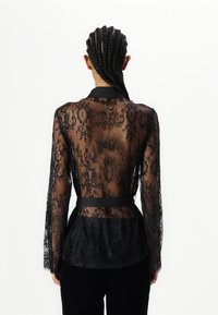 Blusa in pizzo nero trasparente con maniche lunghe, caratterizzata da intricati motivi floreali e una cintura in satin nero in vita. Texture liscia e design elegante.