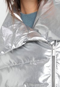 Metaliczna srebrna kurtka puchowa z wysokim kołnierzem, wyposażona w zamek błyskawiczny i teksturowaną tkaninę, prezentująca gładką, błyszczącą powierzchnię.