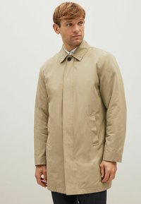 Ung mand iført en beige enkeltradet frakke med krave og sidelommer, som ser til venstre mod en ensfarvet lys baggrund.