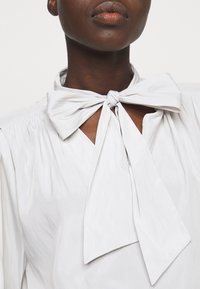 Bílá blůza s hedvábnou texturou, zdobená zavázanou mašlí na výstřihu a výstřihem ve tvaru V, zvýrazněná nařasenými detaily na ramenou.