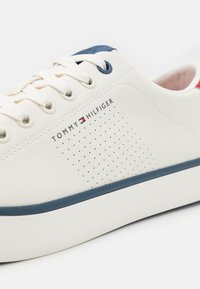 Zapatilla de cuero blanca con acentos en azul marino, punta redonda, detalle perforado en el costado y cordones planos. Presenta un logo en el lateral y una suela texturizada.