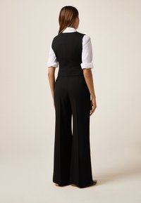 Salopetă neagră, realizată pe măsură, cu picioare largi și un bust ajustat, asortată cu o cămașă albă cu guler. Material neted, design elegant, accente minimale.