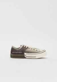 Zapatilla marrón y beige con parte superior texturizada, paneles contrastantes, suela de goma y cierre de cordones. Presenta una marca en el lateral.