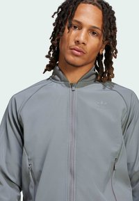 Szary sportowy kurtka z wysokim kołnierzem, gładką fakturą, zamykanymi na zamek kieszeniami i subtelnym logo na piersi. Lekka i dopasowana do sylwetki.