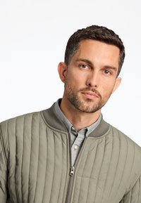 Chaqueta bomber acolchada en color verde oliva con cuello acanalado y cremallera frontal, llevada sobre una camisa de cuello azul claro. Textura suave, diseño moderno.