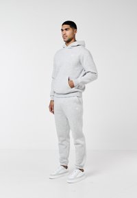 Bărbat purtând hanorac gri deschis și pantaloni sport asortați, cu adidași albi, stând cu o mână în buzunar pe un fundal alb.