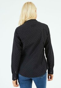 Chemise noire à boutons avec des pois blancs, manches longues et col, présentant une texture de tissu lisse et des poignets à boutons.
