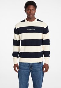 Navy und creme gestreifter Pullover aus Strickstoff mit gerippten Bündchen und Saum, mit dem Logo "GUESS JEANS" auf der Vorderseite. Bequemer Schnitt.