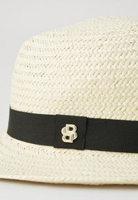 Strohhut mit gewebter Textur, cremefarben, schwarzes Bänder und goldenes Logo-Detail auf der Vorderseite. Rundes Oberteil und breiter Rand.