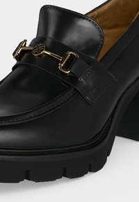 Mocassin en cuir noir avec détail en métal doré en mors de cheval et talon épais, présentant des coutures lisses et une doublure intérieure couleur tabac.