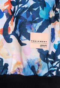 Tela de estampado floral colorido con tonos de azul, rosa y verde, con un dobladillo acanalado en negro y una etiqueta rosa que dice "EQUIPMENT adidas."