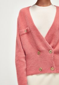 Cardigan rose en peluche avec un design en V, doté de boutons dorés et d'une petite poche poitrine. Tricot texturé au fini doux.