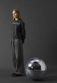 Grå sweatshirt med texten "CHROME", kombinerad med löst sittande mörka jeans. En stor, rund silverfärgad discokula reflekterar ljuset i förgrunden.